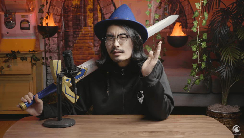 Tim Lyu dons a wizard had and brandishes the Master Sword from the Legend of Zelda video game series as he explains the history of isekai anime in front of a backdrop designed to look like a fantasy dungeon in a scene from the Anime Explained Youtube video essay series .